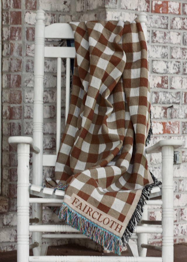 Checkered blanket with 'Faircloth' branding draped over a white rocking chair against a brick wall.