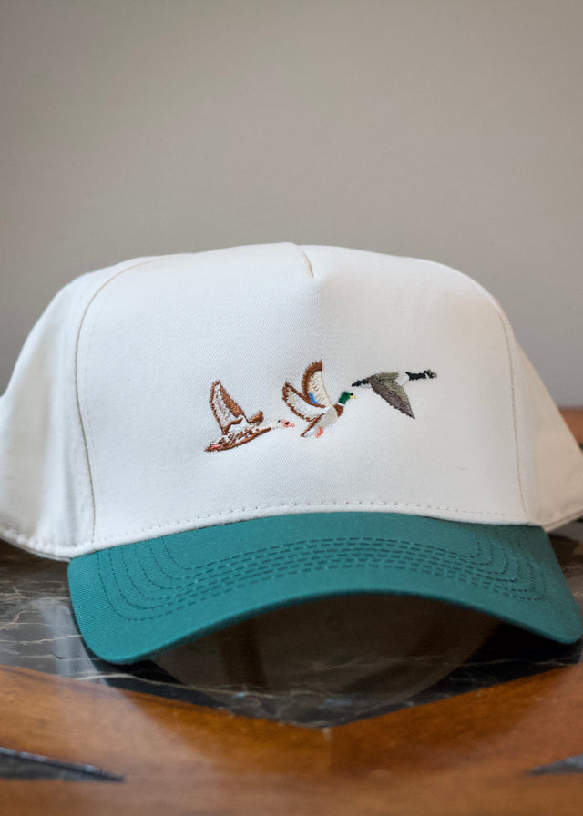 White cap with green bill featuring embroidered two ducks and a goose on a wooden surface.