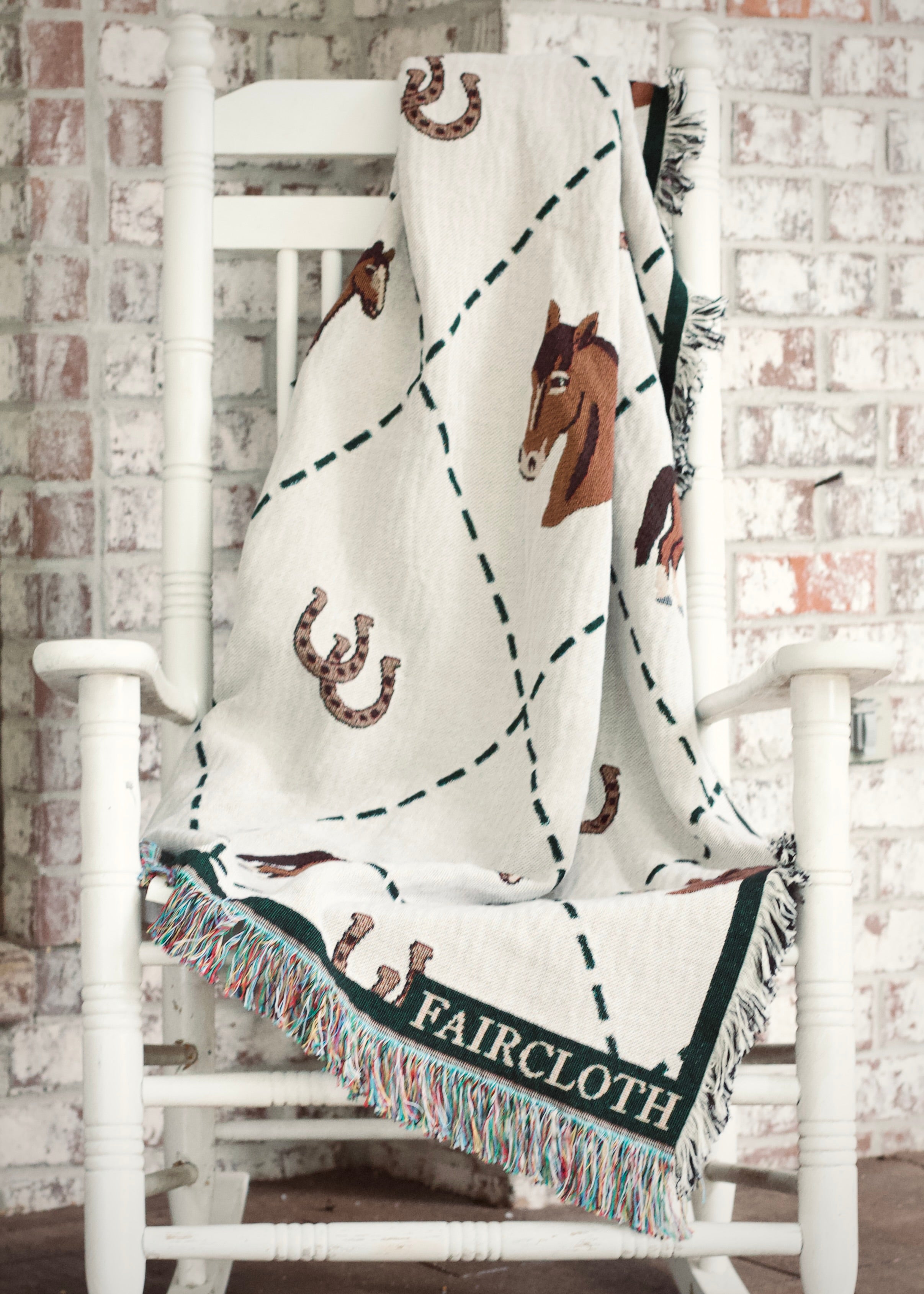 Blanket with horse and horseshoe patterns draped over a white chair against a brick wall.