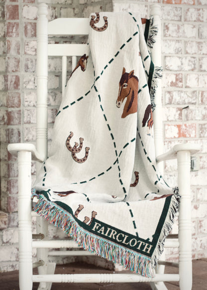 Blanket with horse and horseshoe patterns draped over a white chair against a brick wall.