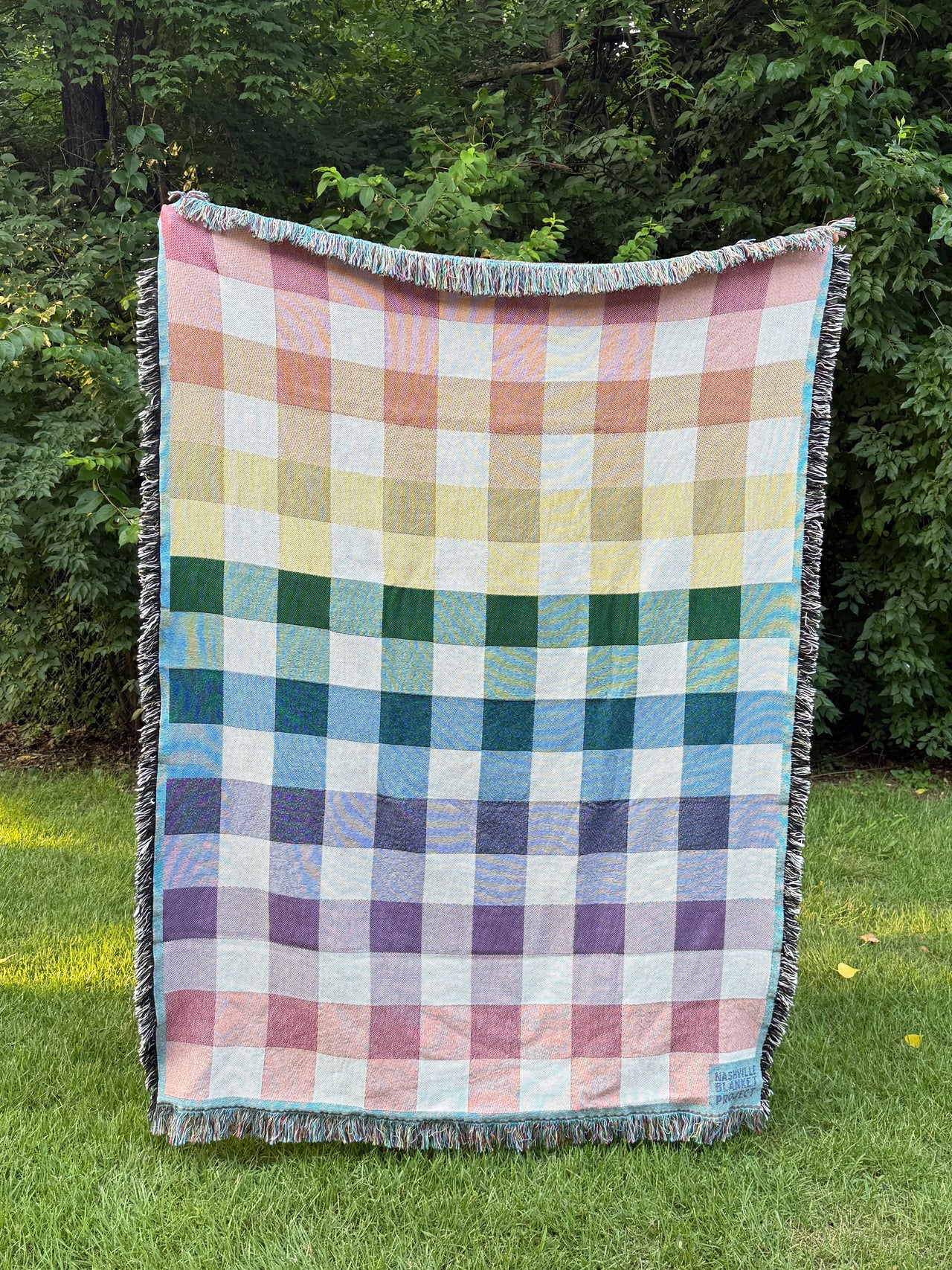 rainbow gingham blanket on grass with trees in the background